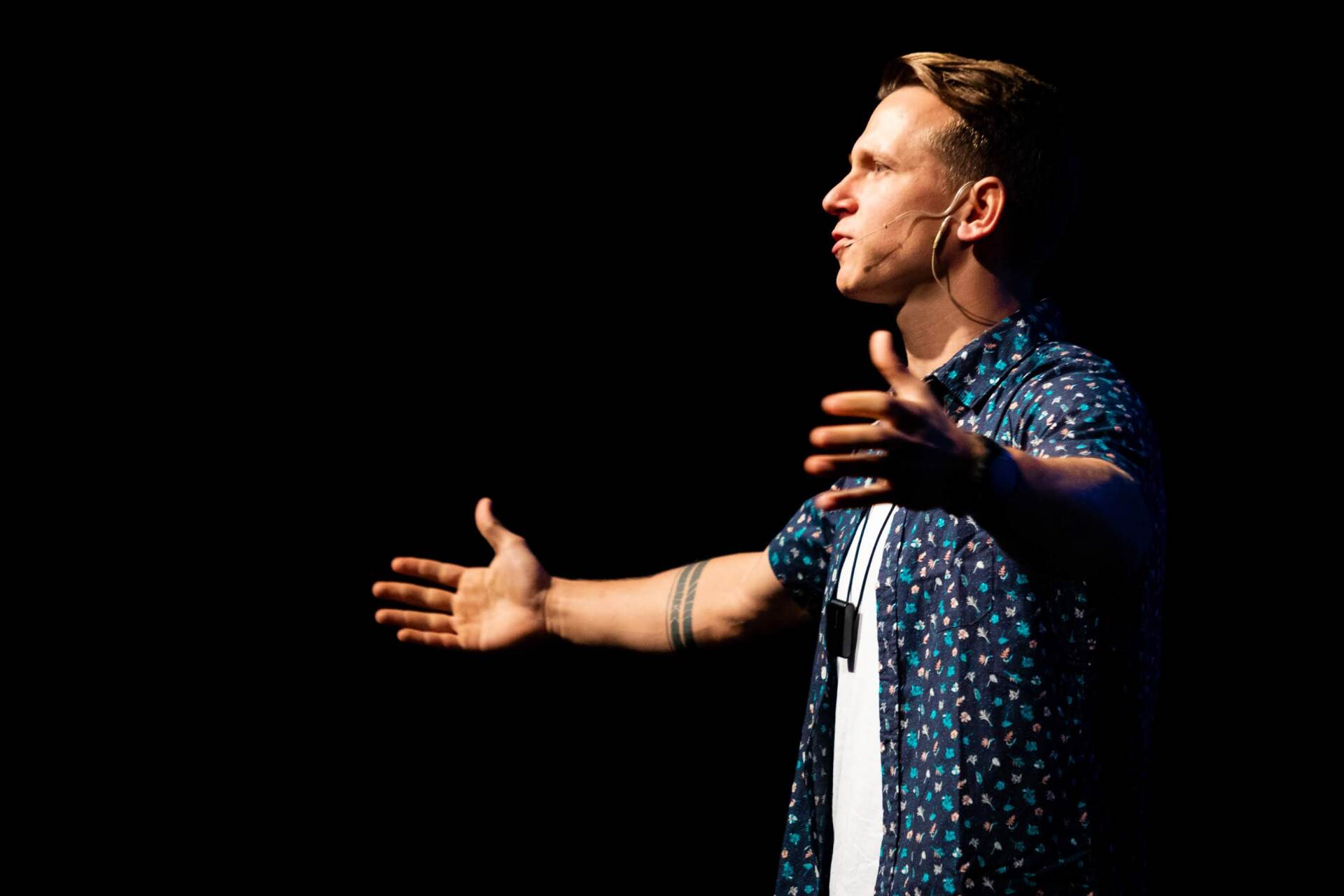 A man in a blue shirt is standing in the dark with his arms outstretched.