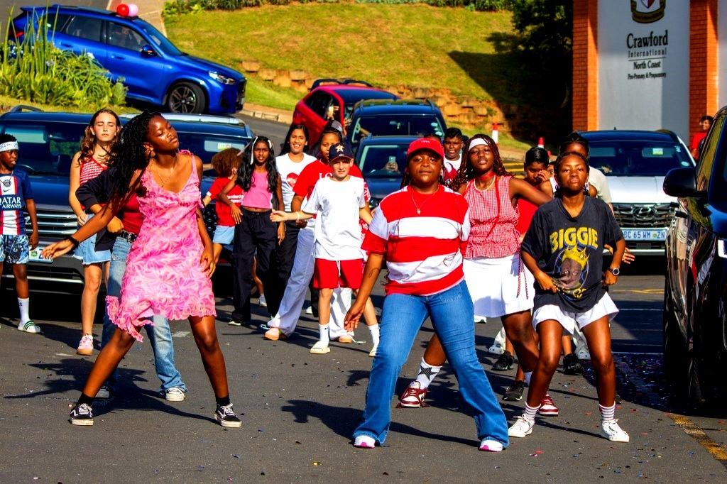 Group of people dancing in the street with cars in the background; sunny day.