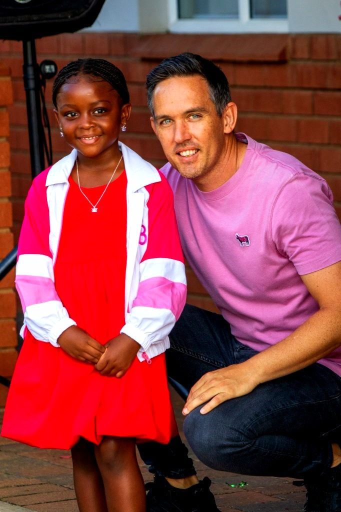 A man kneels next to a child, both smiling in front of a brick wall. The child wears a red dress and jacket.