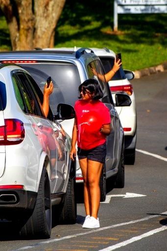 Woman in red shirt and black shorts stands between two white SUVs, while two people in the SUVs hold up phones.