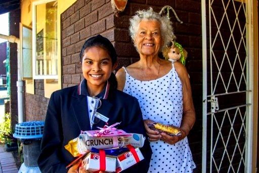 Girl in school uniform giving gifts to an older woman in front of a house.