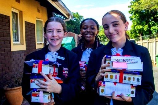 Three students holding stacked gifts, smiling, in school uniforms.