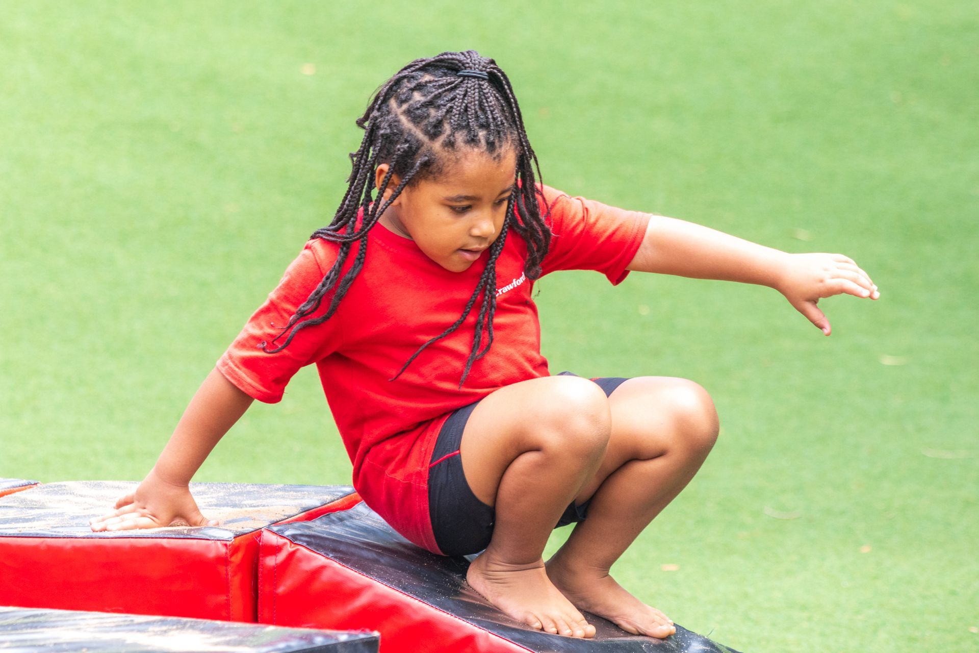 Child in red shirt and black shorts squats on a red block, balancing with arms outstretched.