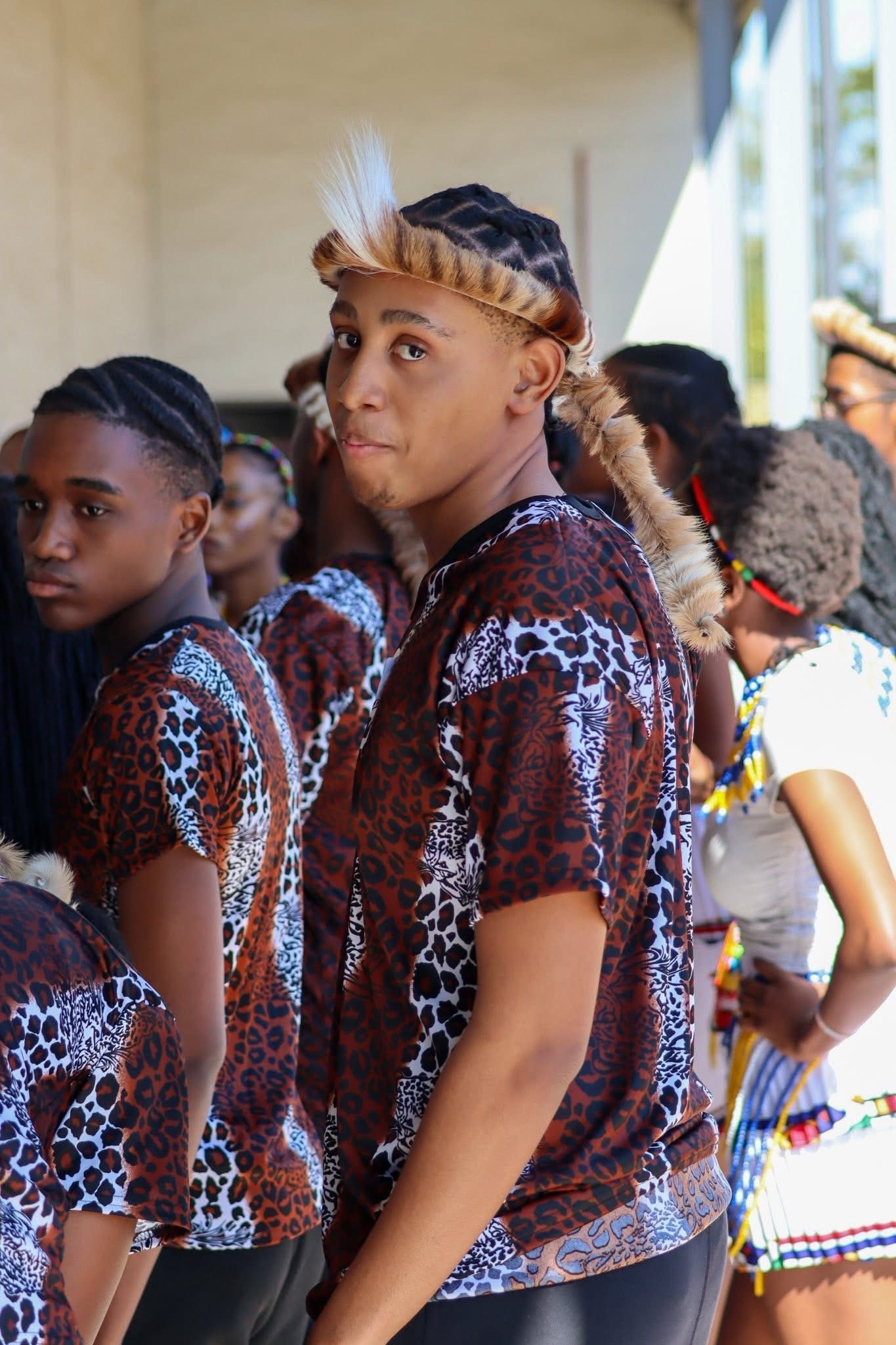 Group of people in matching patterned shirts, one with braided hair and feather accessory.