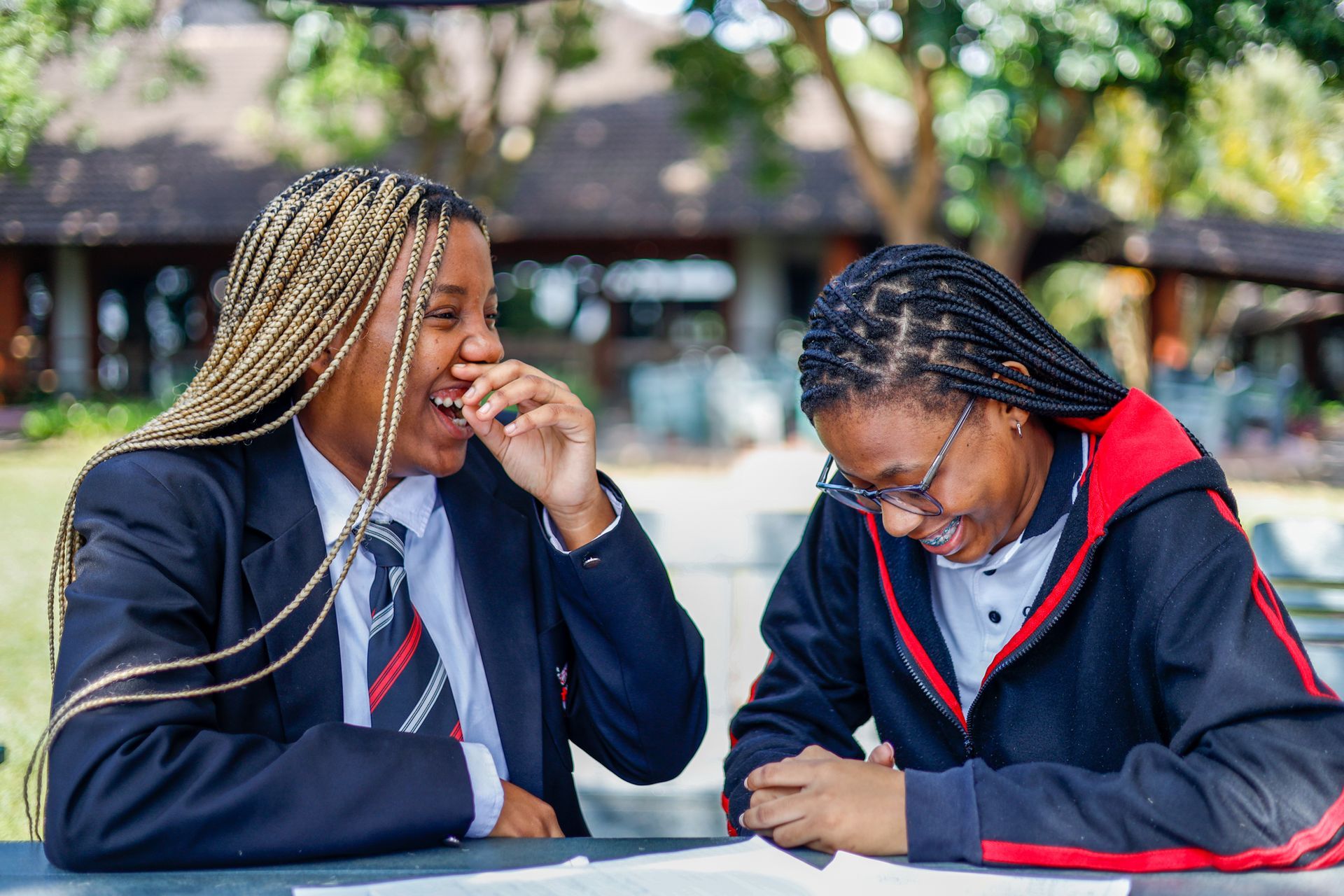 Two students laugh while sitting at a table outdoors. One has blonde braids, the other glasses.