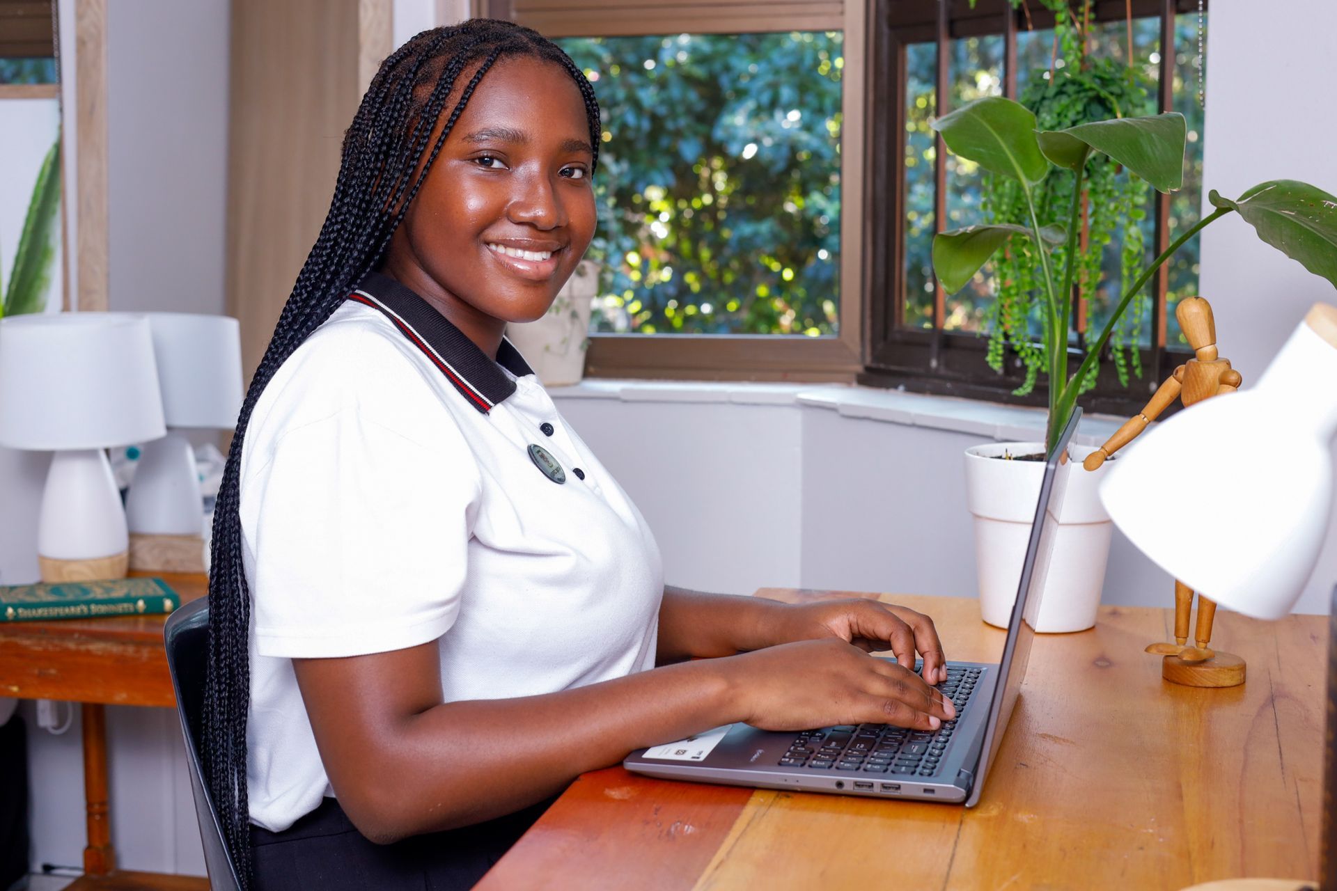 Woman with braided hair, smiling, typing on a laptop at a wooden desk near a window.