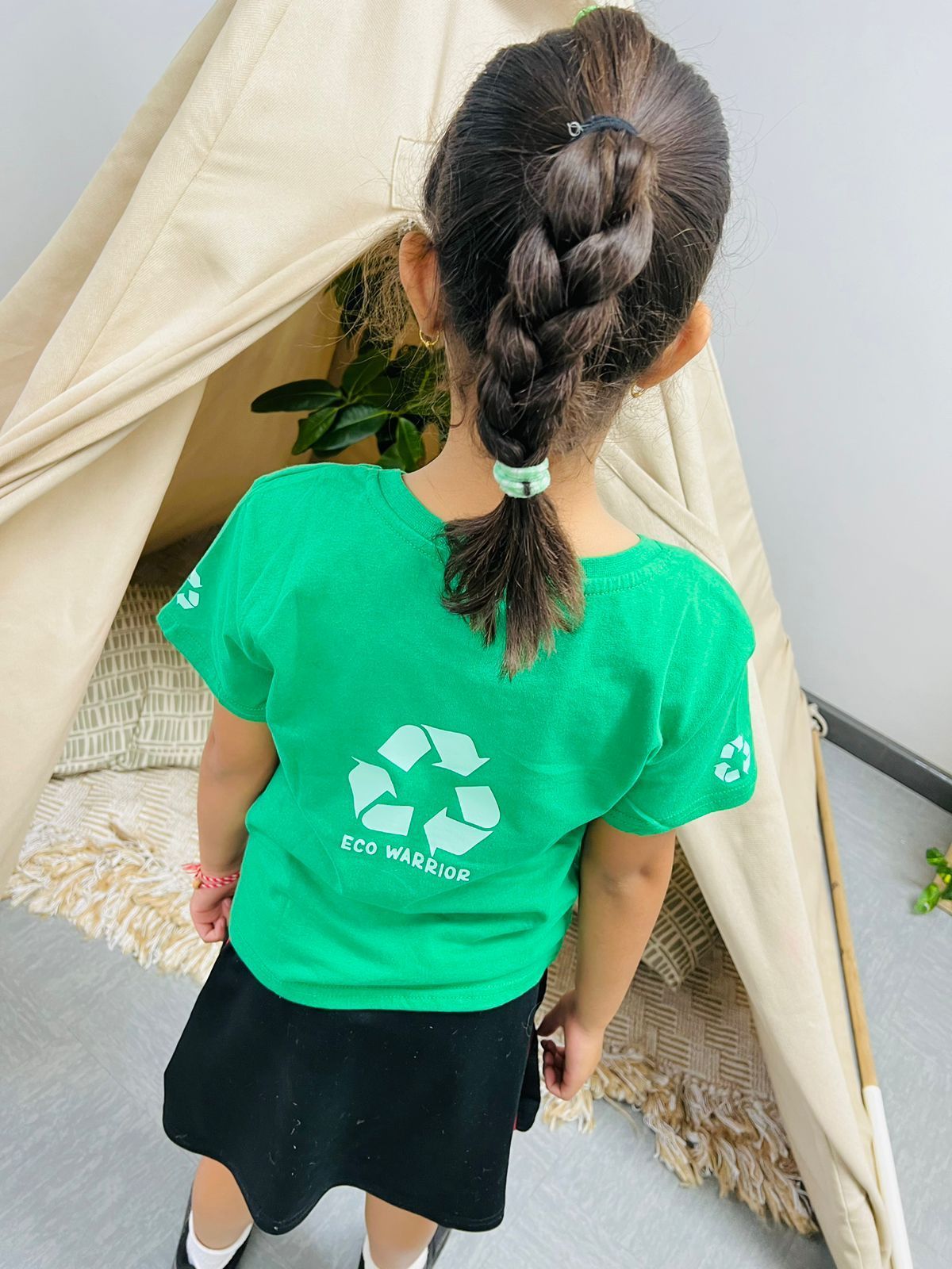 A little girl wearing a green t-shirt with a recycling symbol on it is standing in front of a teepee.