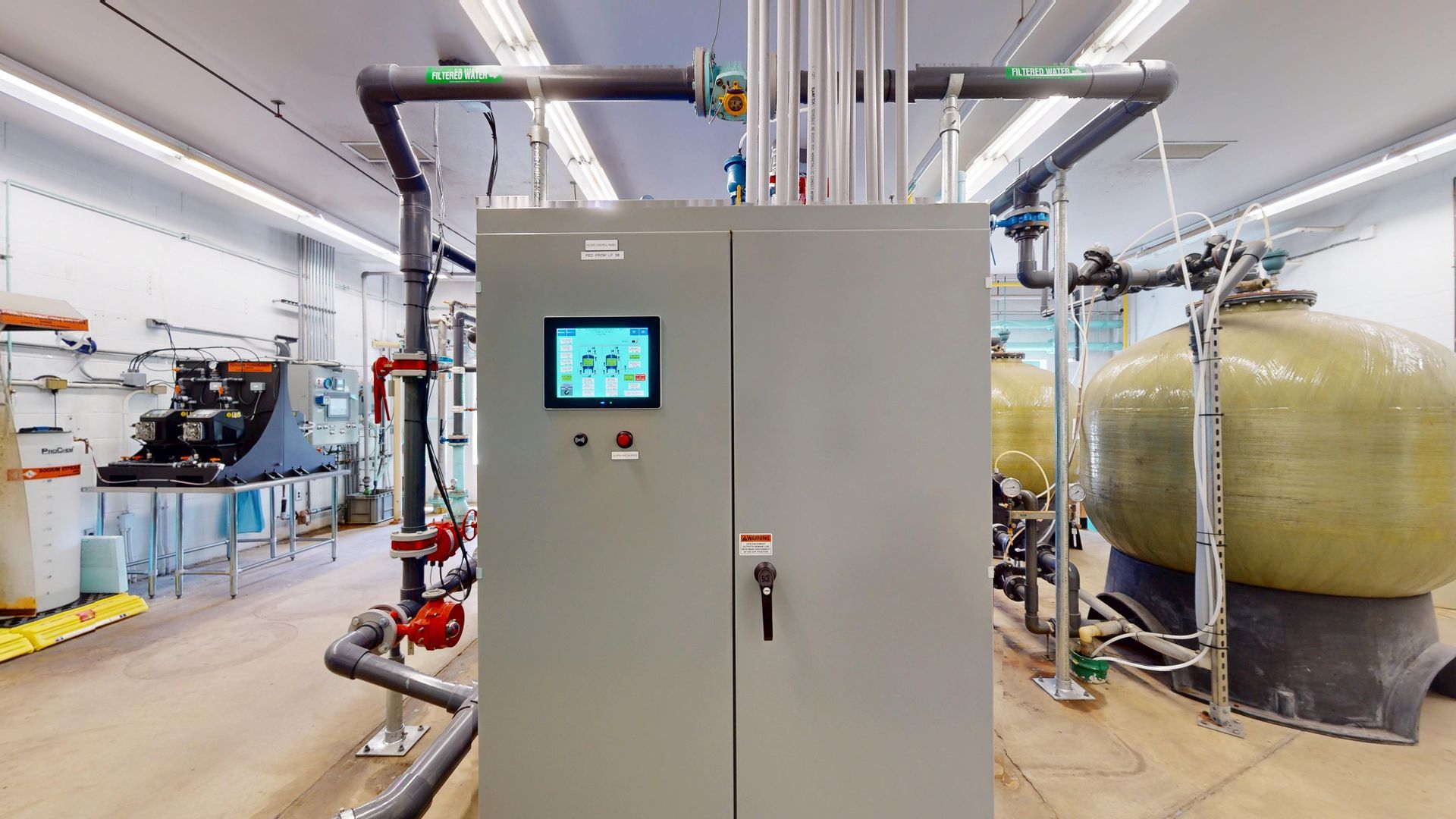 Control panel in a utility room with pipes, valves, and tanks; industrial setting.