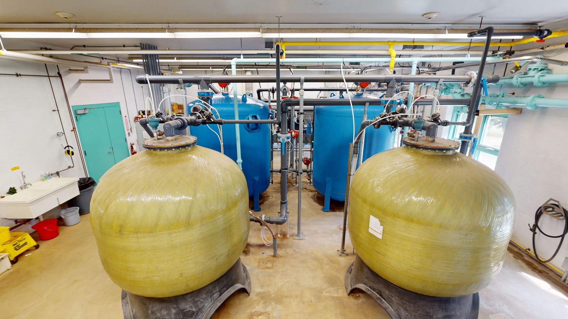 Water filtration system in a utility room with large yellow and blue tanks, pipes, and other equipment.
