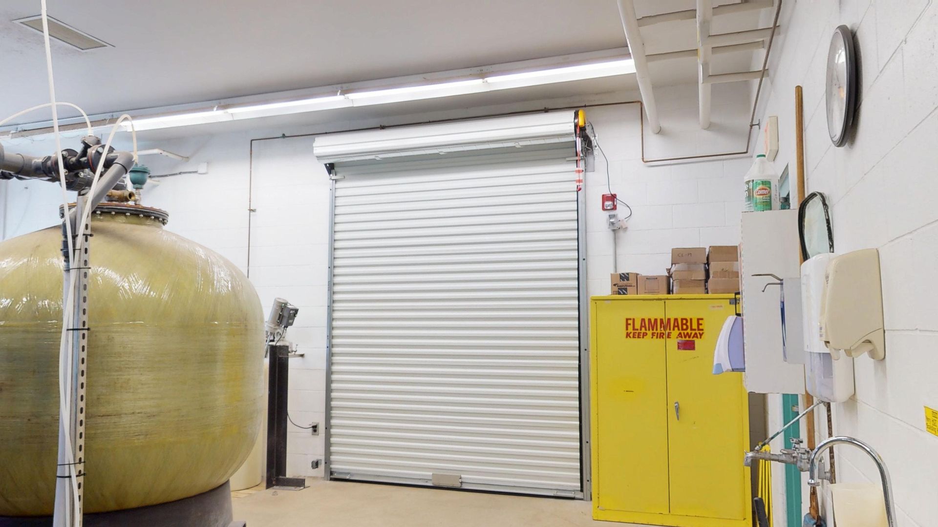 A utility room with a large tank, a closed roll-up door, and a yellow flammable storage cabinet.