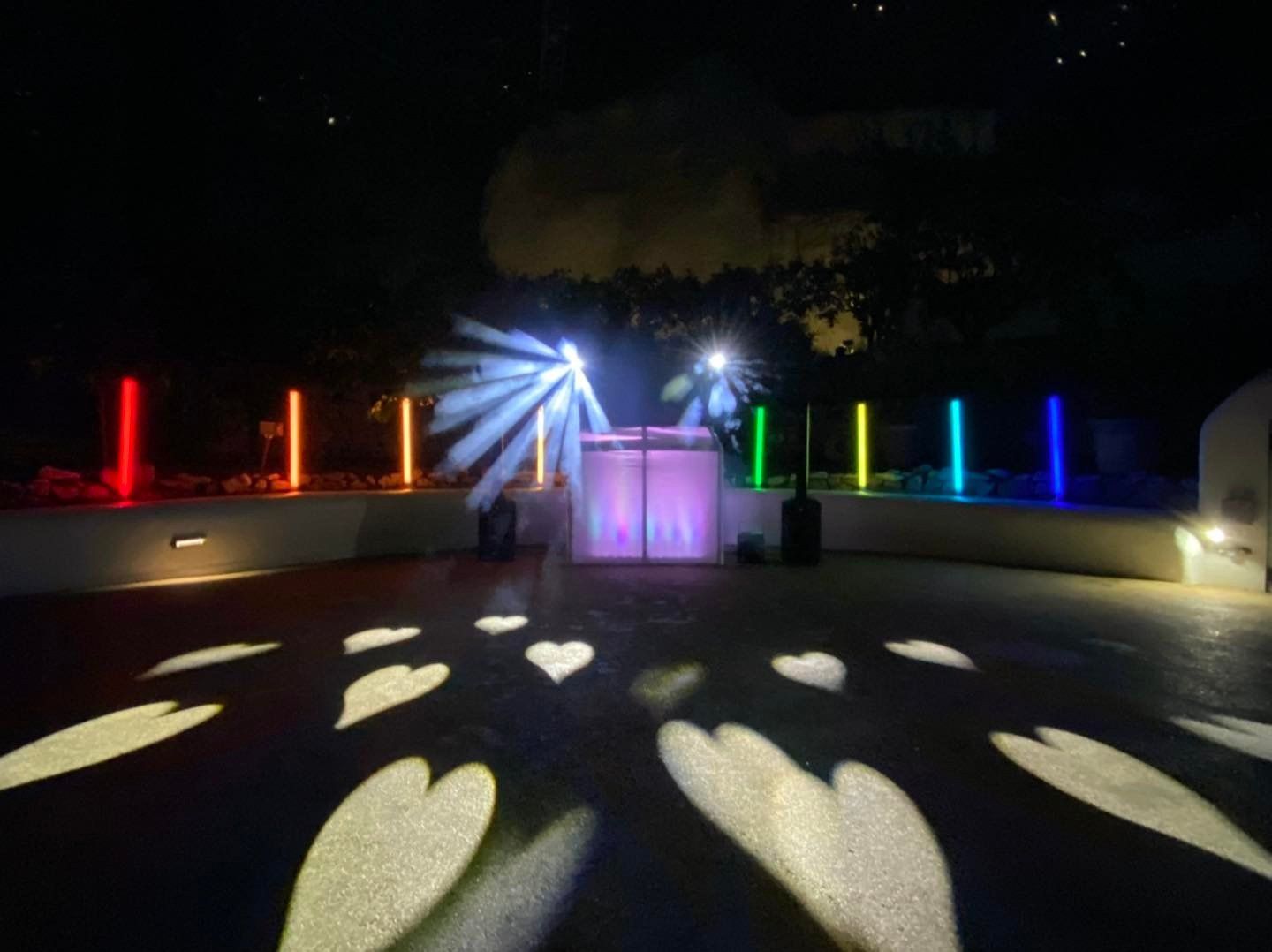 Nighttime outdoor event with DJ setup, colorful lights, and heart-shaped projections on the ground.