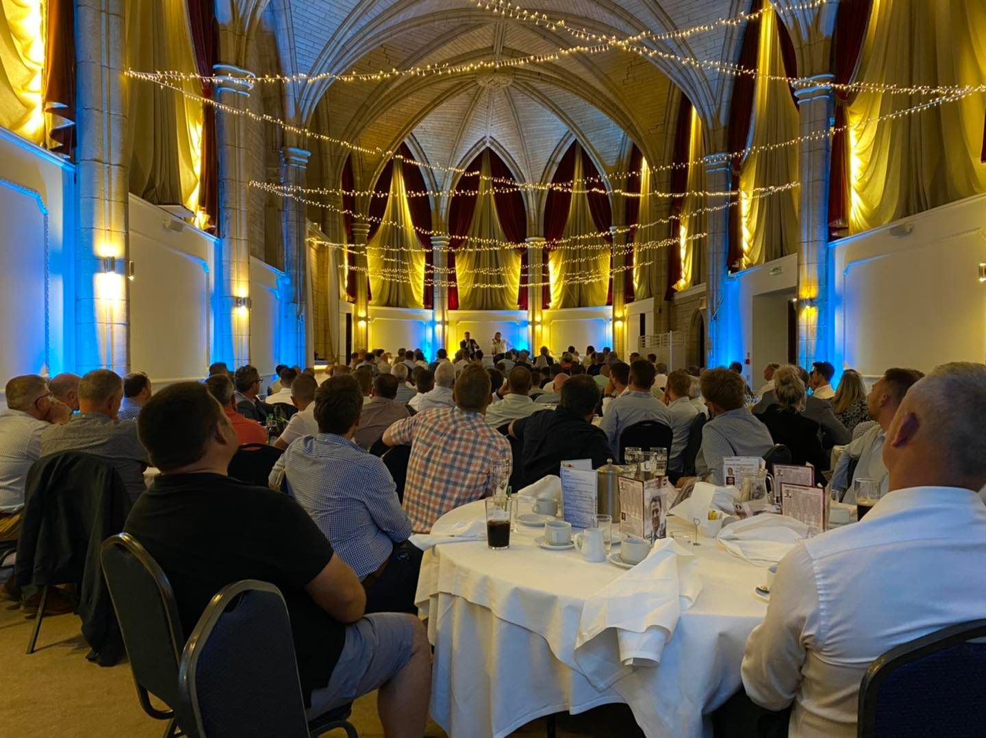 A banquet hall with guests seated at round tables, listening to a presentation at the front, under ambient lighting.