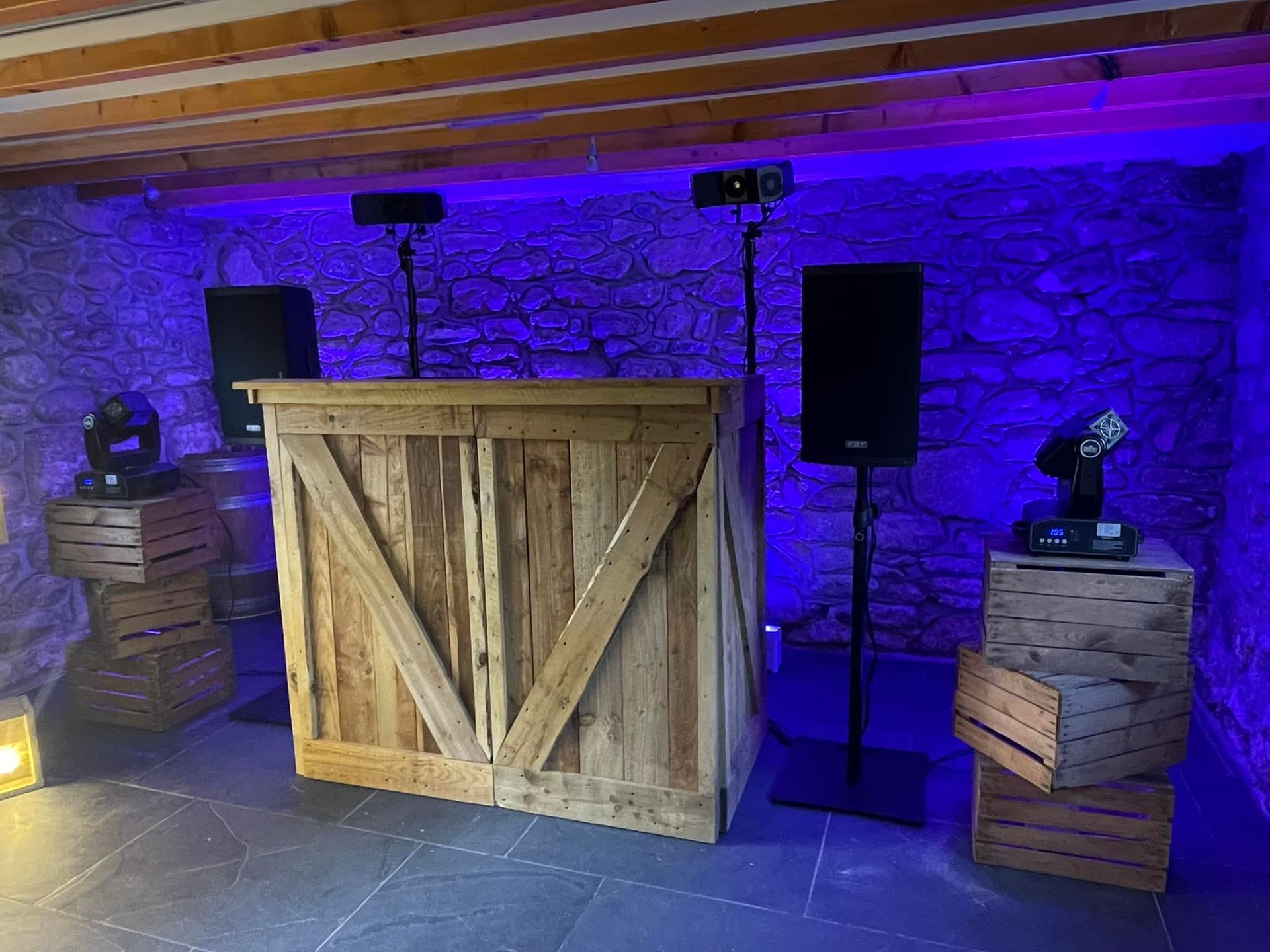 DJ setup with wood bar, speakers, and lights against a stone wall lit with blue.