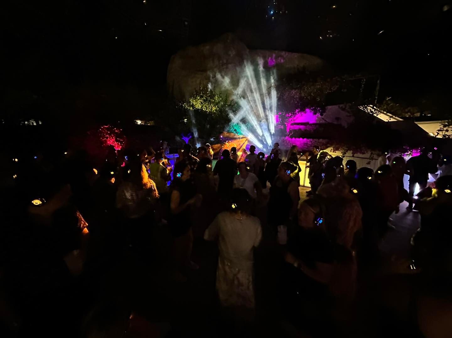 Nighttime crowd at a party with colorful lights, rock formation backdrop.