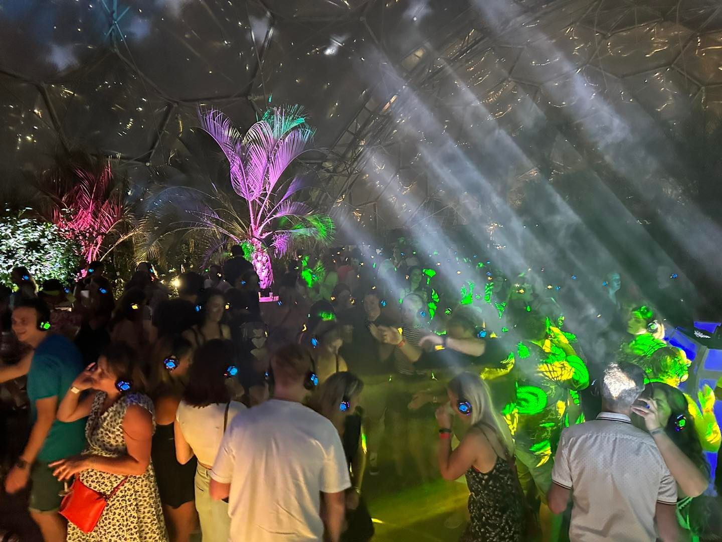 People dancing at a silent disco under a dome. Green and blue lights with people wearing headphones.