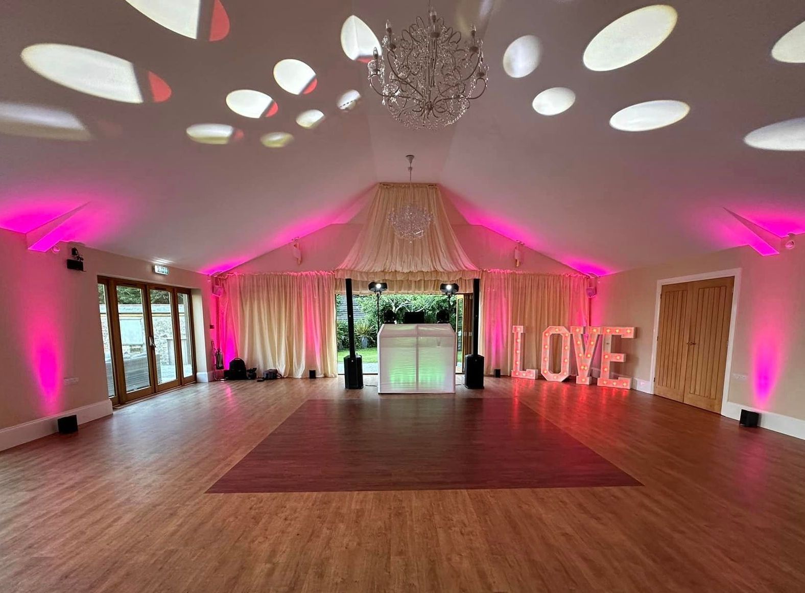 Interior of a wedding reception hall with dance floor, pink lighting, LOVE letters, and a DJ booth.