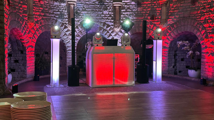 DJ setup with red and white lights in a stone archway. Dance floor in front, stacks of plates to the left.