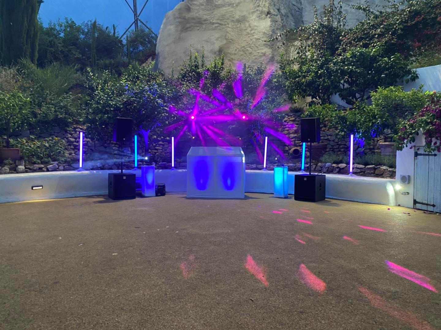 Outdoor DJ setup with purple and blue lighting against greenery.