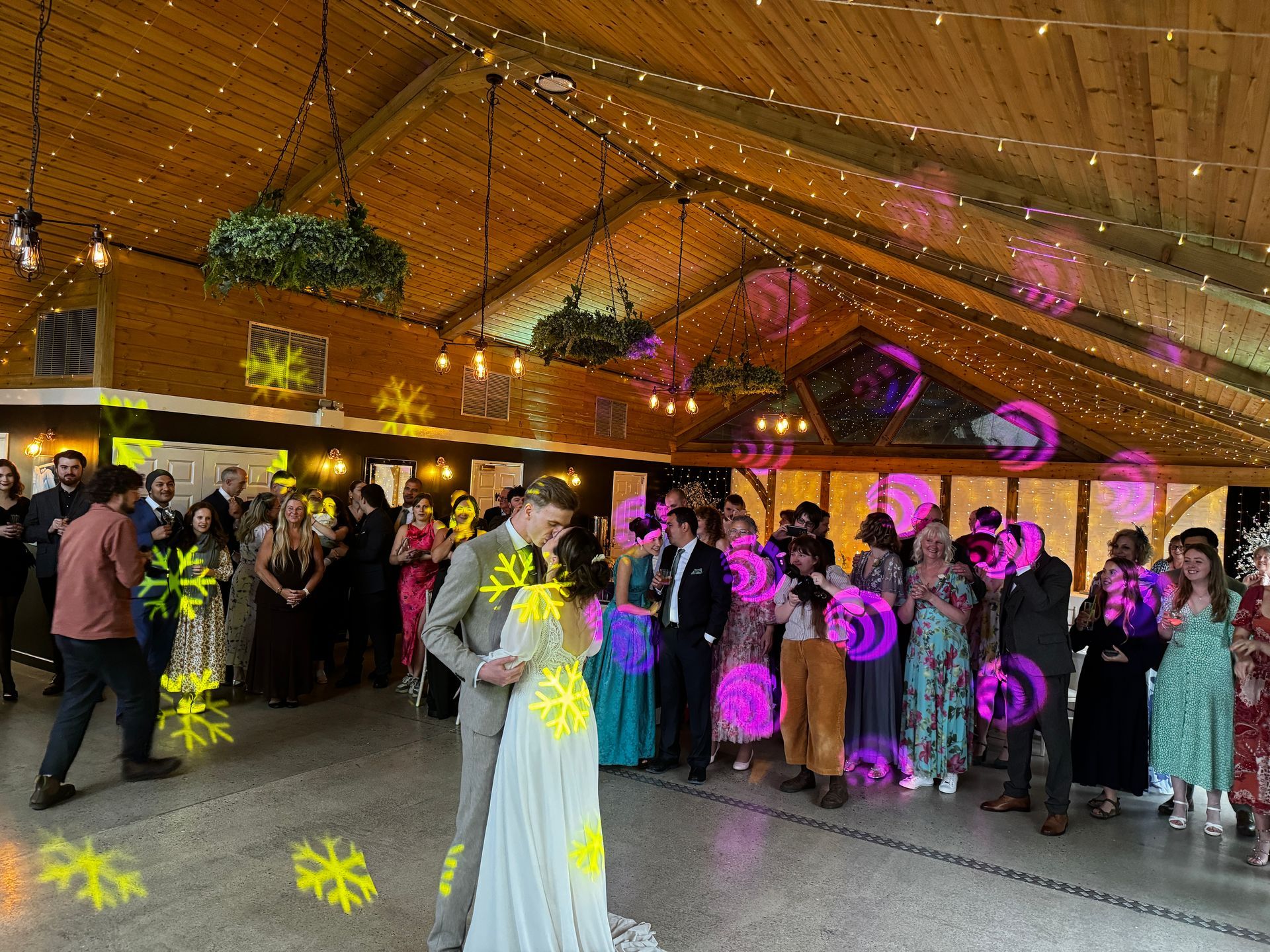 Wedding reception, couple dancing. Guests watch. Interior with string lights, wood ceiling.