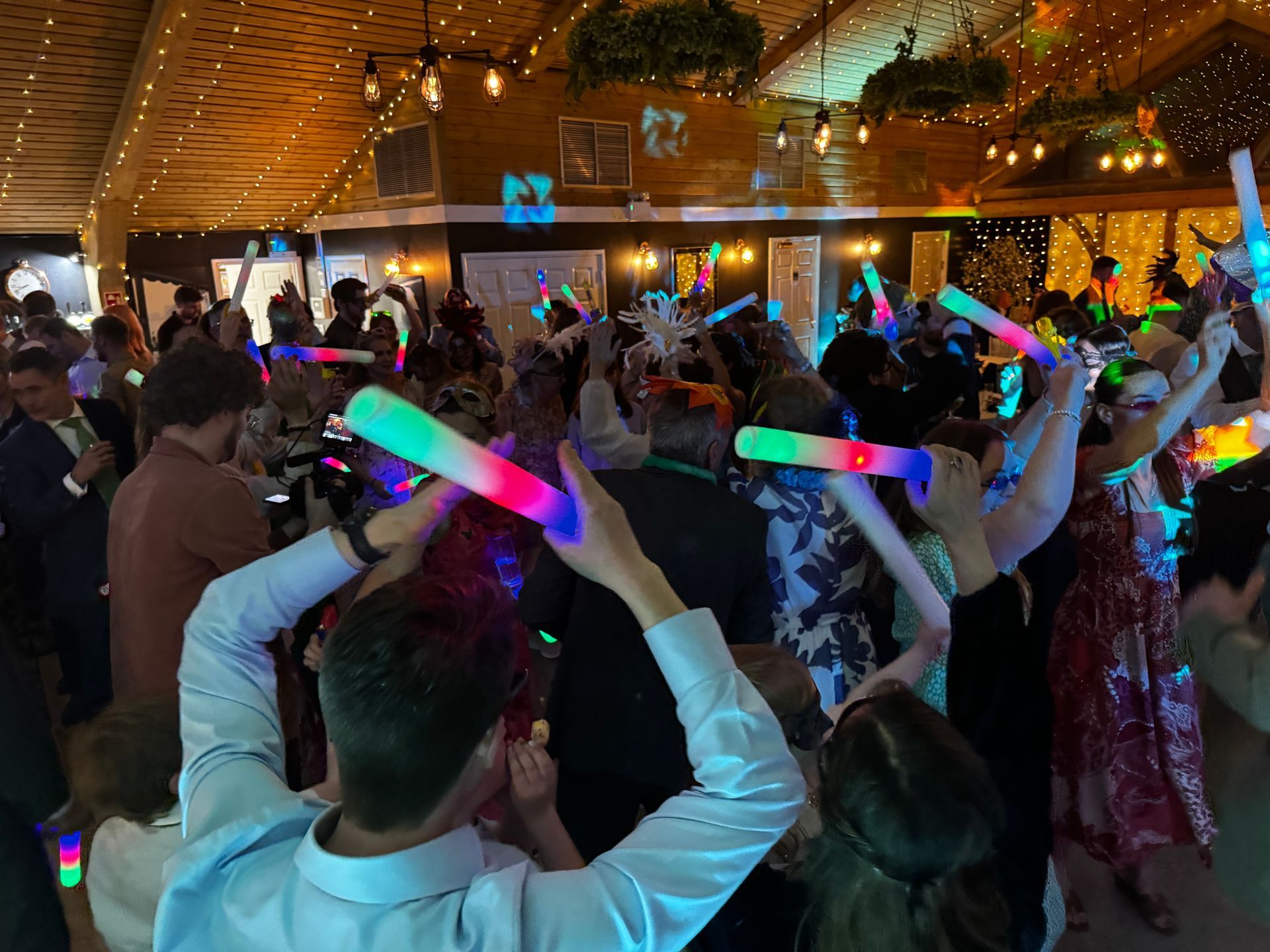 People dancing at an event, holding colorful light-up wands. Indoors with string lights and decor.
