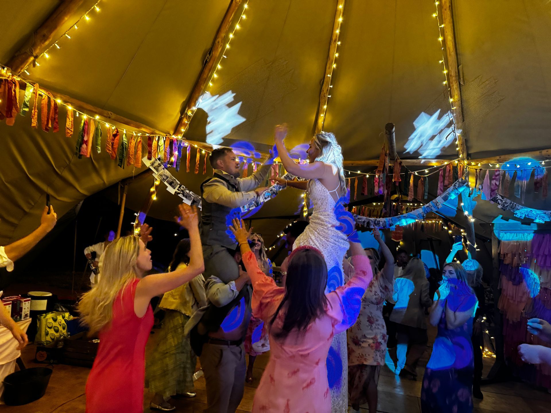 Wedding reception inside a decorated teepee. Guests lift the bride and groom up in the air, celebrating.