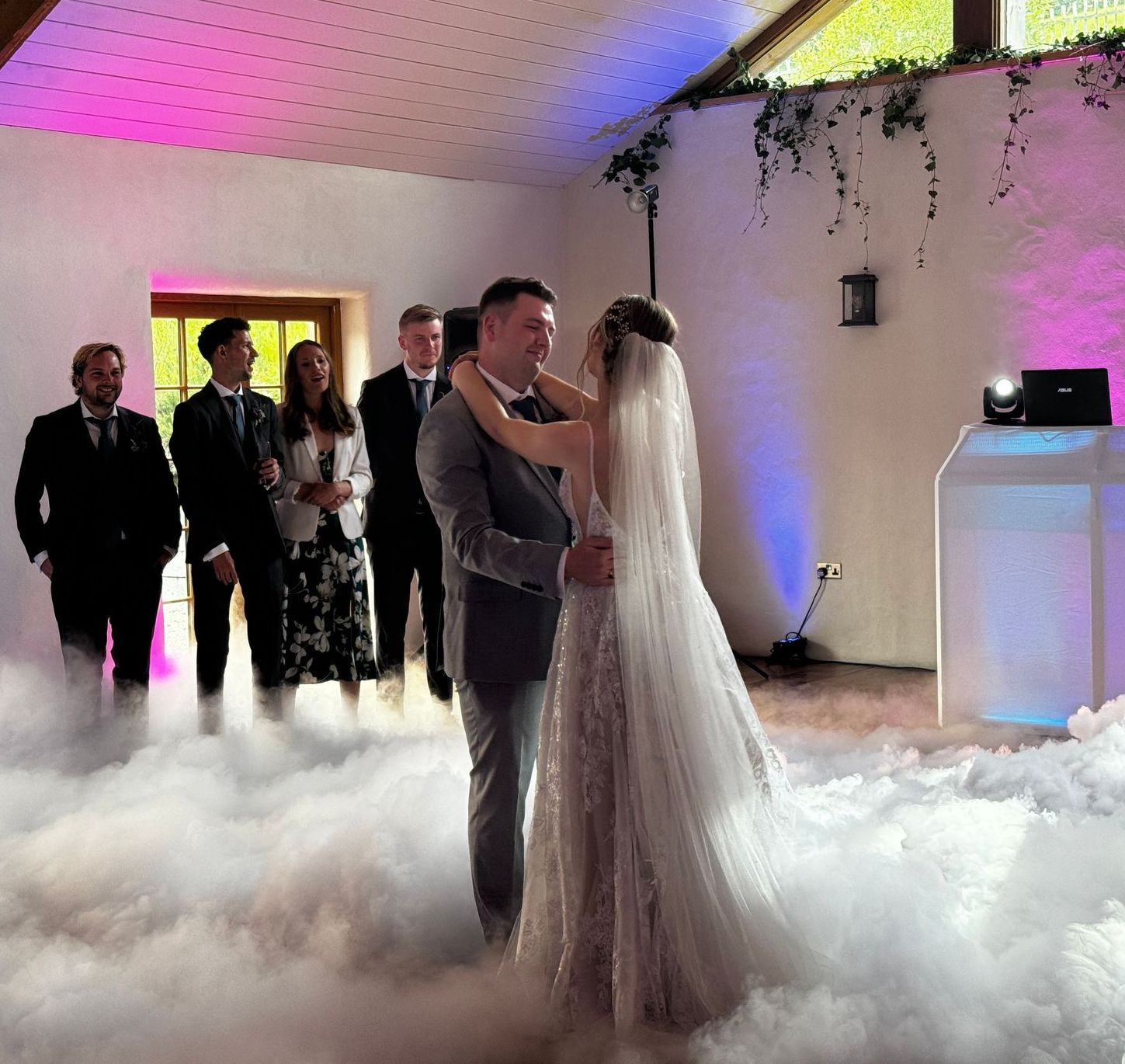 Bride and groom dance in a cloud of fog at their wedding reception, guests watch.