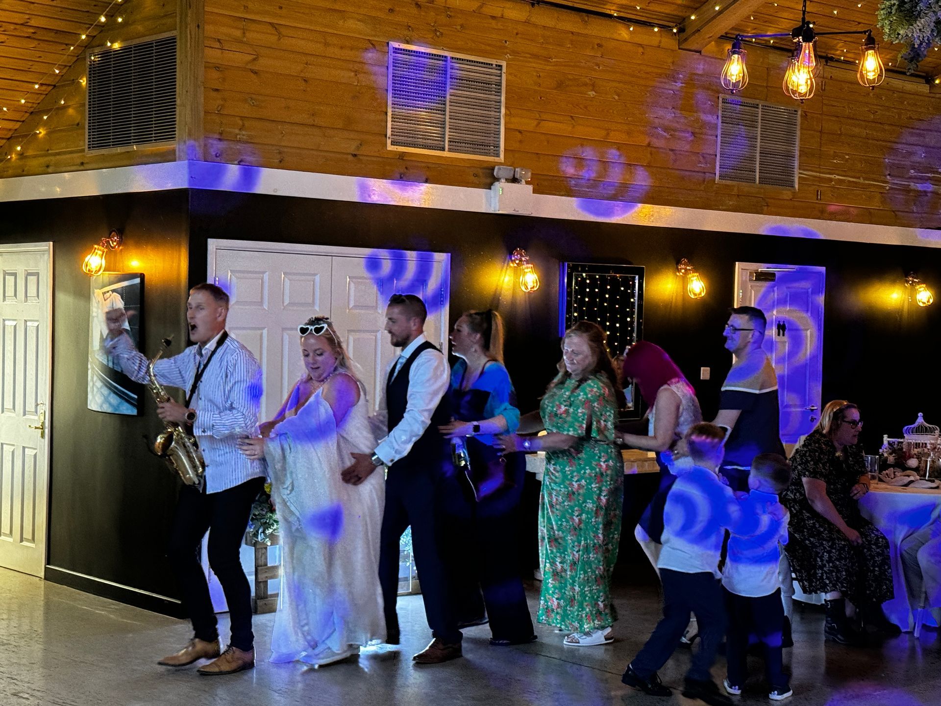 People dancing at a wedding reception, indoors. Bride & groom in center, others in formal wear. Purple lighting.