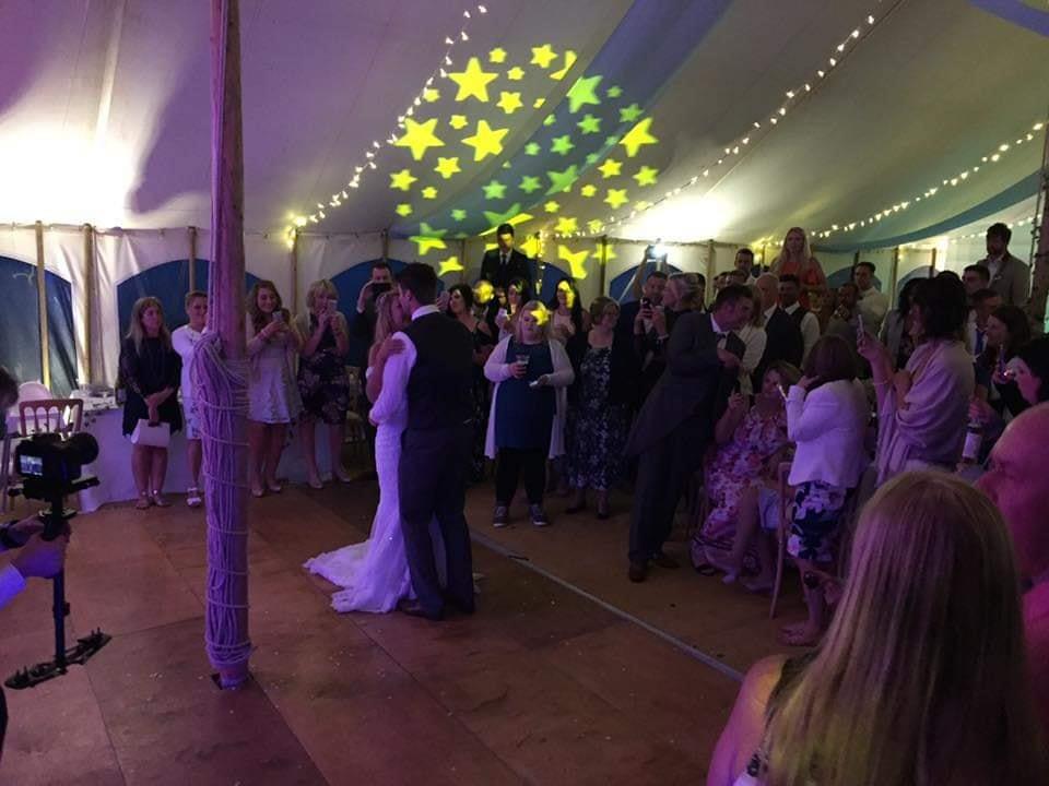 Bride and groom share a dance in a tent, guests watch, lights project stars on the ceiling.