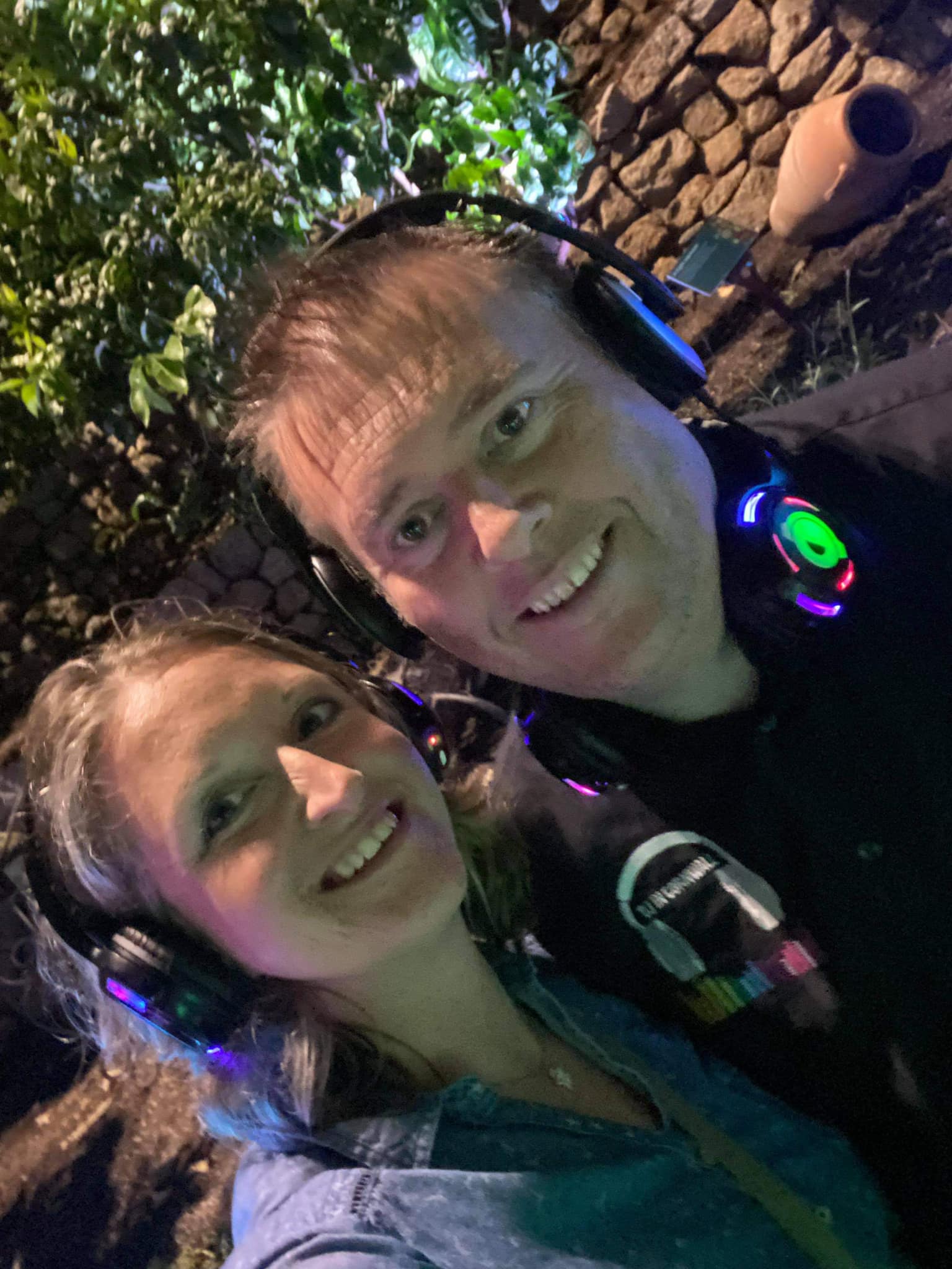 Smiling couple wearing headphones at a silent disco, posing for a selfie at night, with stone background.