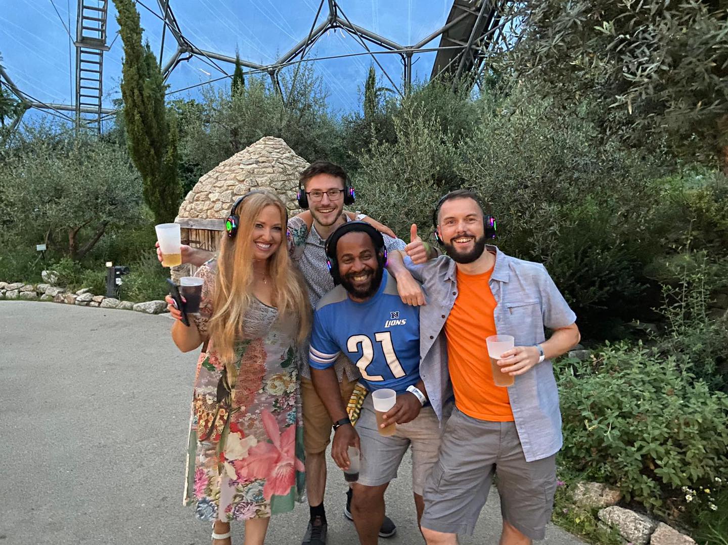 Five people smiling, wearing headphones, holding drinks, in front of greenery and a domed structure.