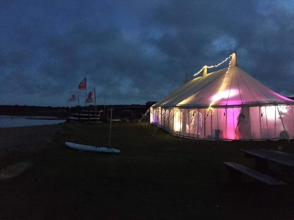A clear tent glows with pink and white lights at dusk, beside a body of water and a small sailboat.