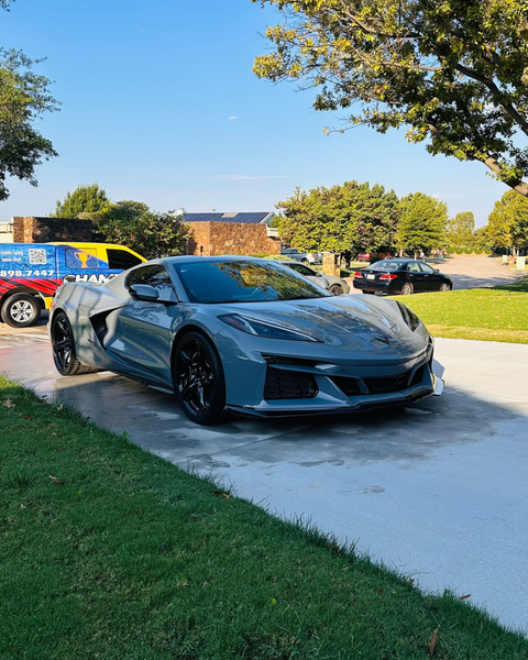 Gray sports car parked on a sunny driveway beside a colorful pickup truck and lakeside trees.