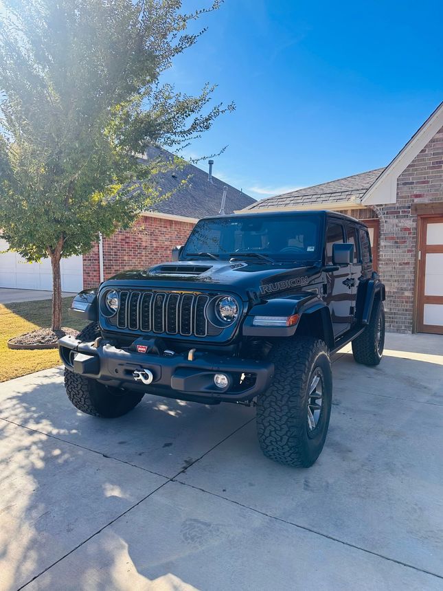 Black Jeep Wrangler parked in a sunny driveway beside a house and tree.