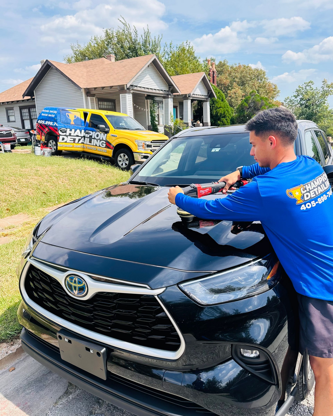 Man polishing a black Toyota SUV in a driveway, with a house and work truck in the background