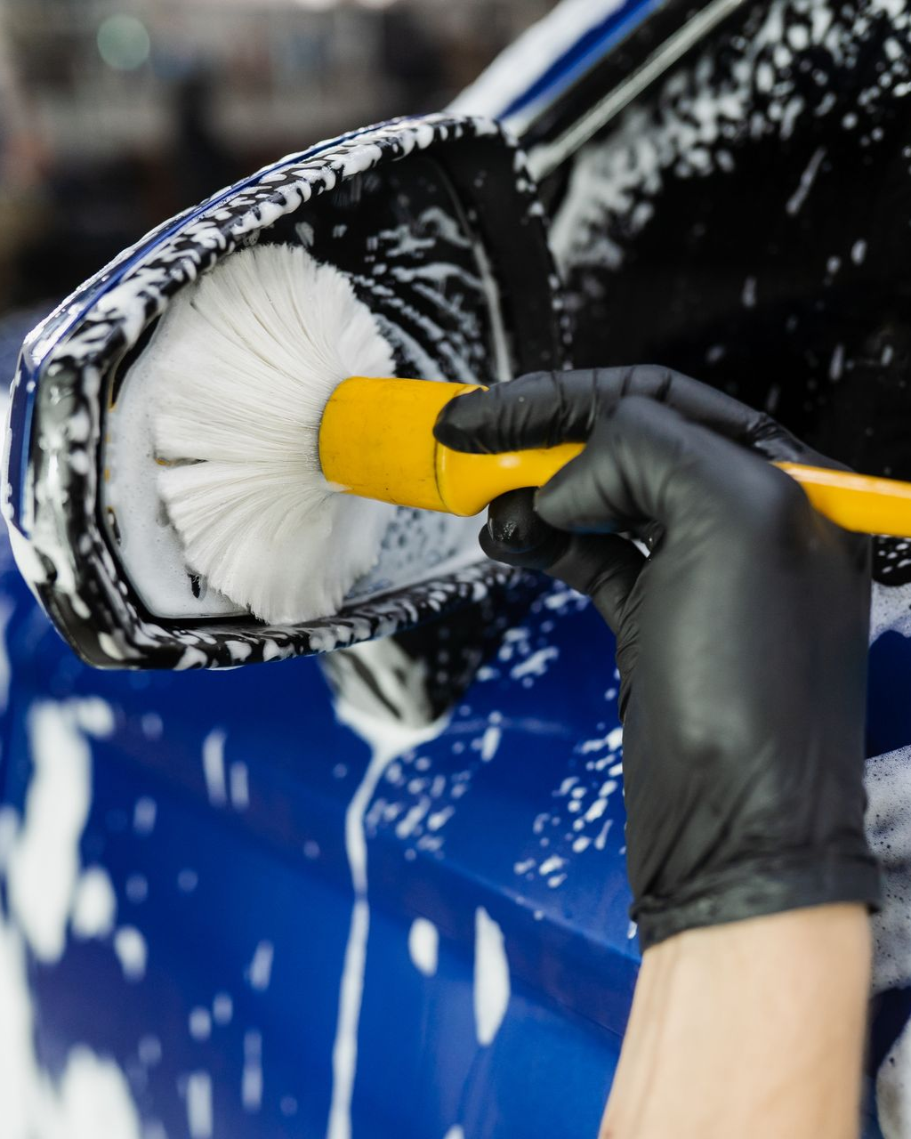 Gloved hand washing a blue car’s side mirror with a white brush and soap foam