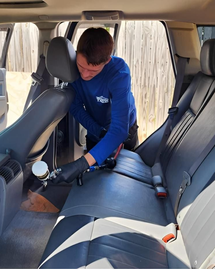 Person cleaning the back seat of a car with a vacuum, wearing a blue shirt and black gloves