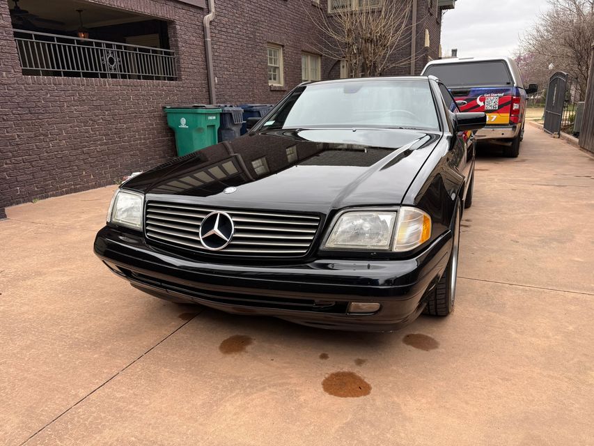 Black Mercedes sedan parked on a driveway beside a brick building, with another car behind it.