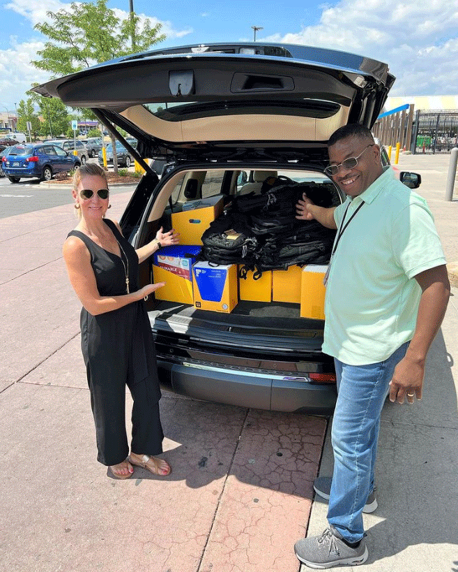 Two people stand by an SUV's open trunk filled with supplies, likely for donation.