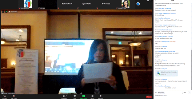 Woman reading from paper in front of screen, in a meeting room, online participants visible.