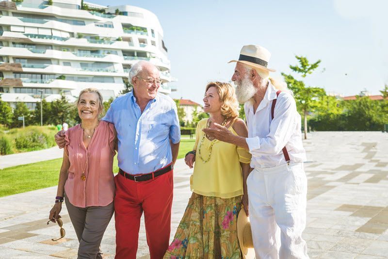 Four smiling seniors walking outdoors, near a building with green space, talking, and enjoying the sun.