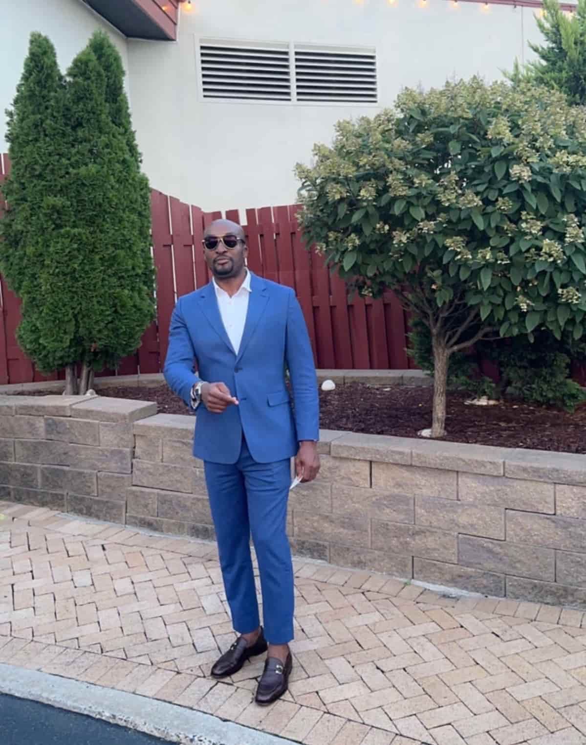 A person in a blue suit, white shirt, and dark sunglasses stands on a brick patio in front of a stone wall and trees.