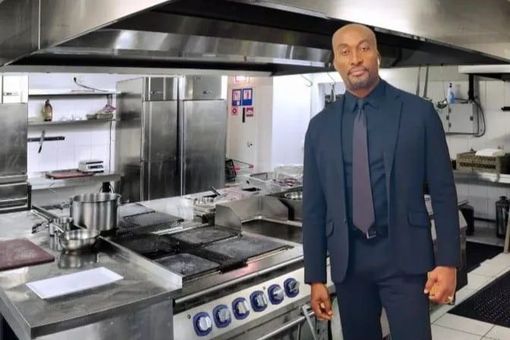 Man in a suit stands in a commercial kitchen with stainless steel appliances.
