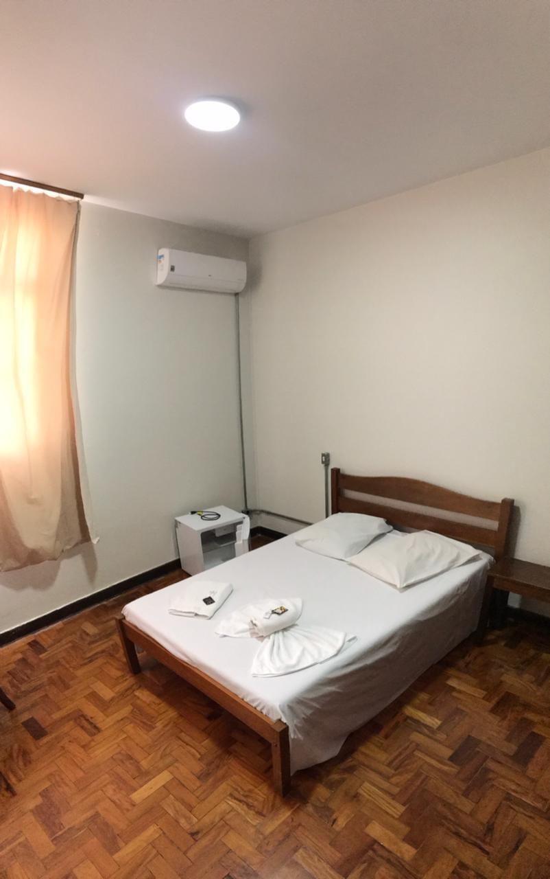 Quarto de hotel pequeno com cama de casal, mesa de cabeceira, ar condicionado e piso de parquet de madeira.