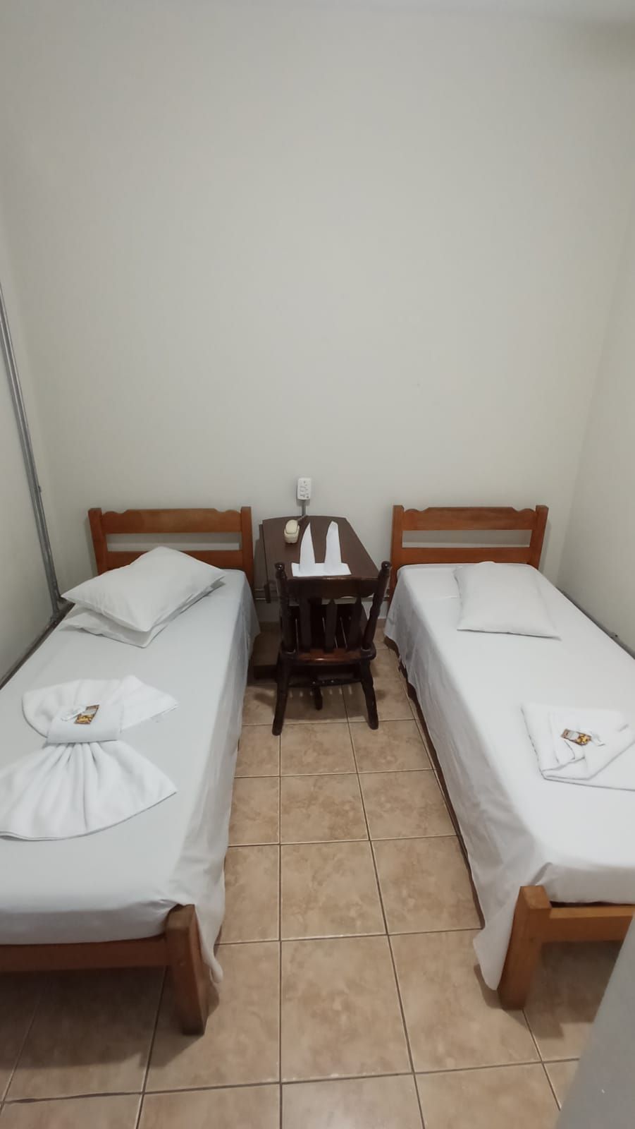 Duas camas de solteiro, uma mesinha e uma cadeira em um quarto de hotel simples.