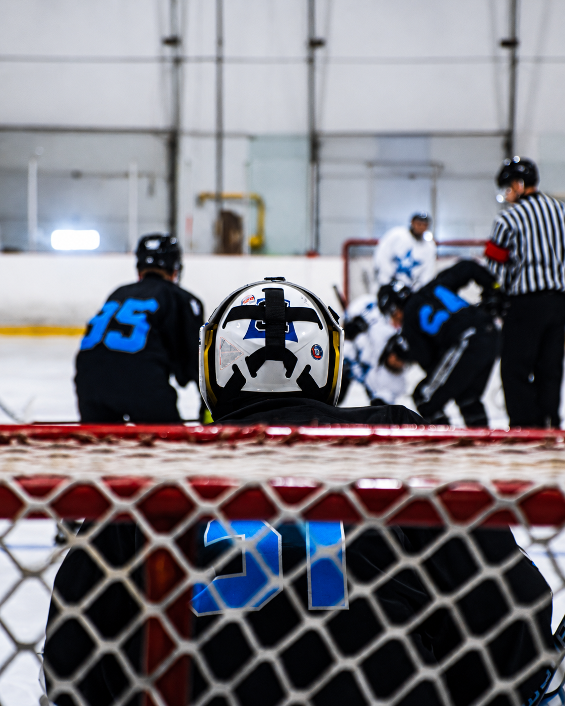 Ice hockey goalie in black and blue, seen from behind through the net during a faceoff.