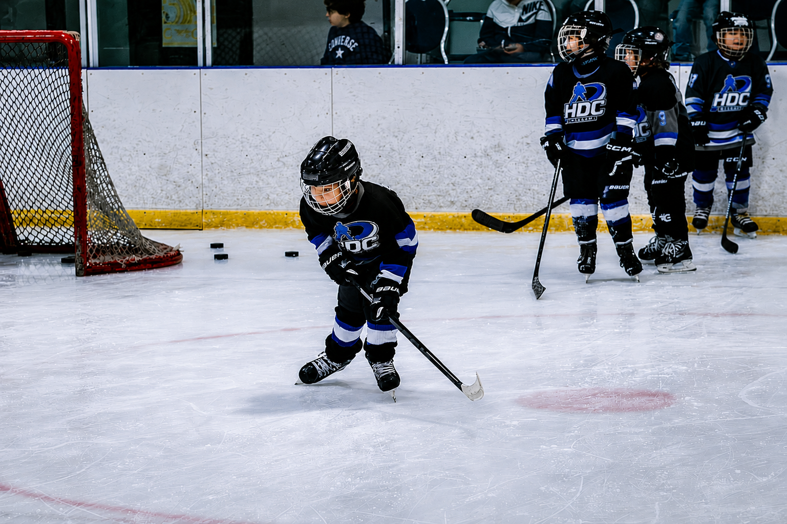 Youth hockey players on ice near the goal, one skating with the puck while teammates stand behind him.