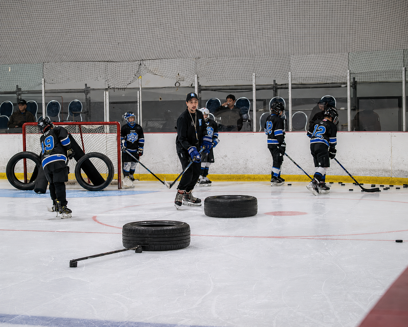 Youth hockey players in black jerseys practice on ice with tires and sticks as obstacles.