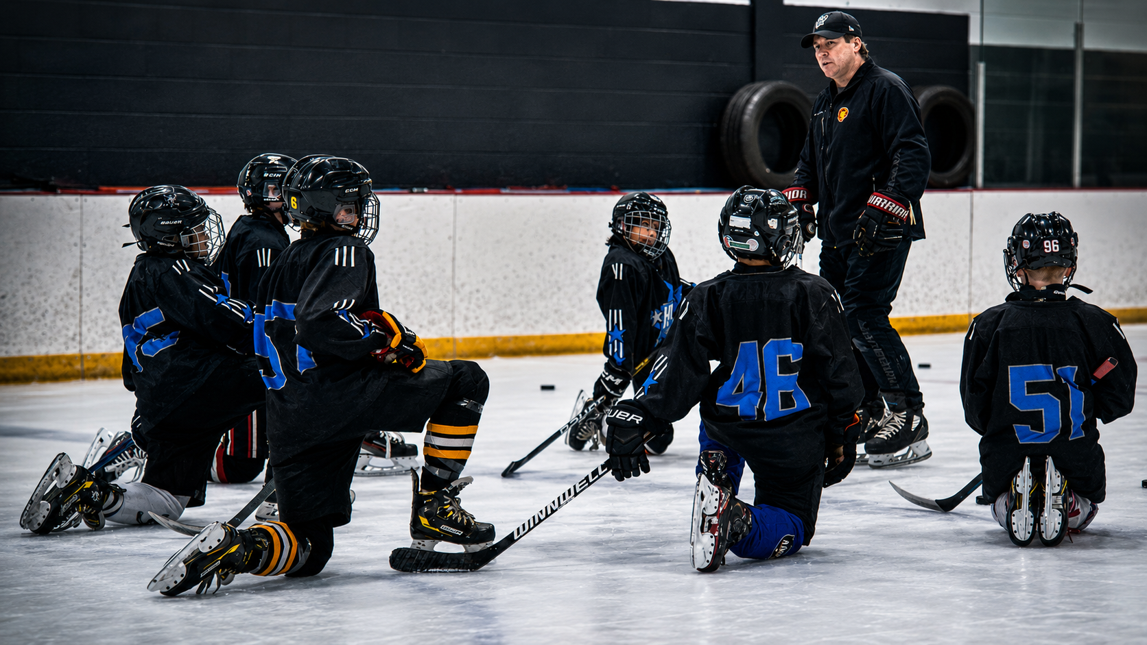 Youth hockey coach instructing kneeling players on an indoor ice rink, with blue jerseys and black helmets.