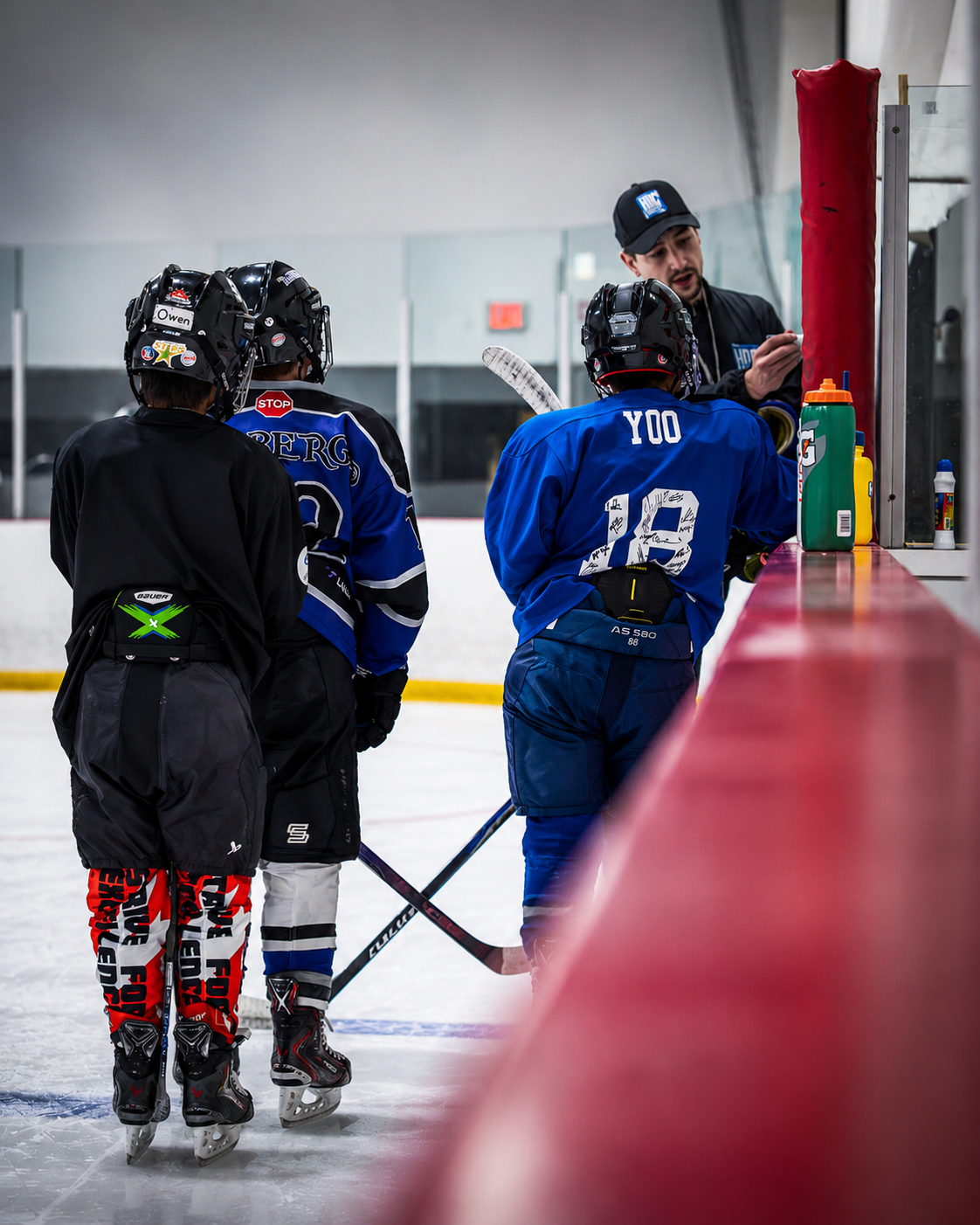Youth hockey players in blue jerseys gather at rink boards during practice, with a coach nearby.