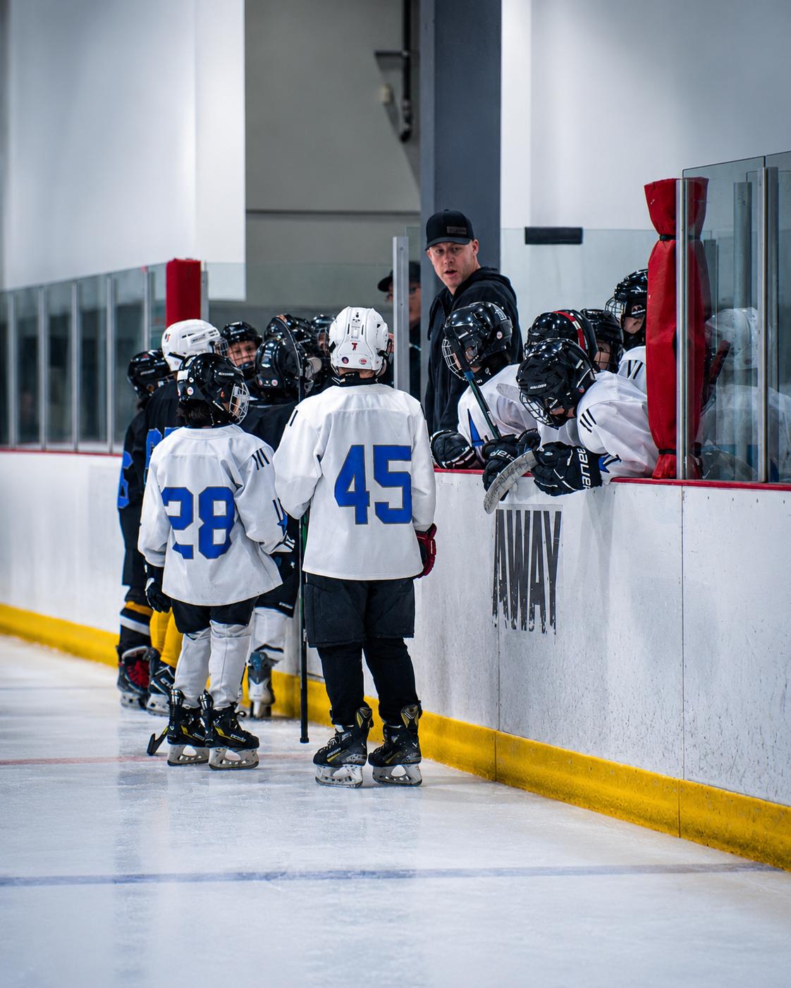 Youth hockey players in white jerseys huddle at the rink boards with a coach nearby.